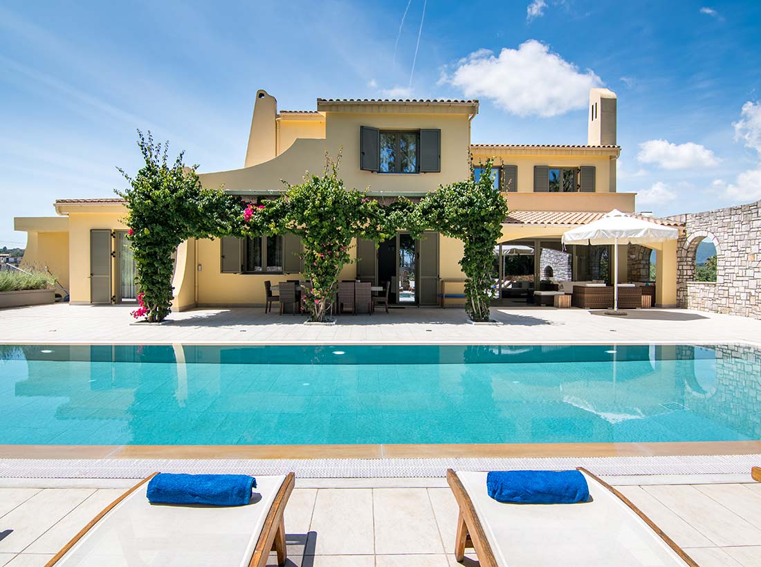 Villa Alpha, an ochre two-storey house with bougainvillea, seen across its pool with loungers on the terrace