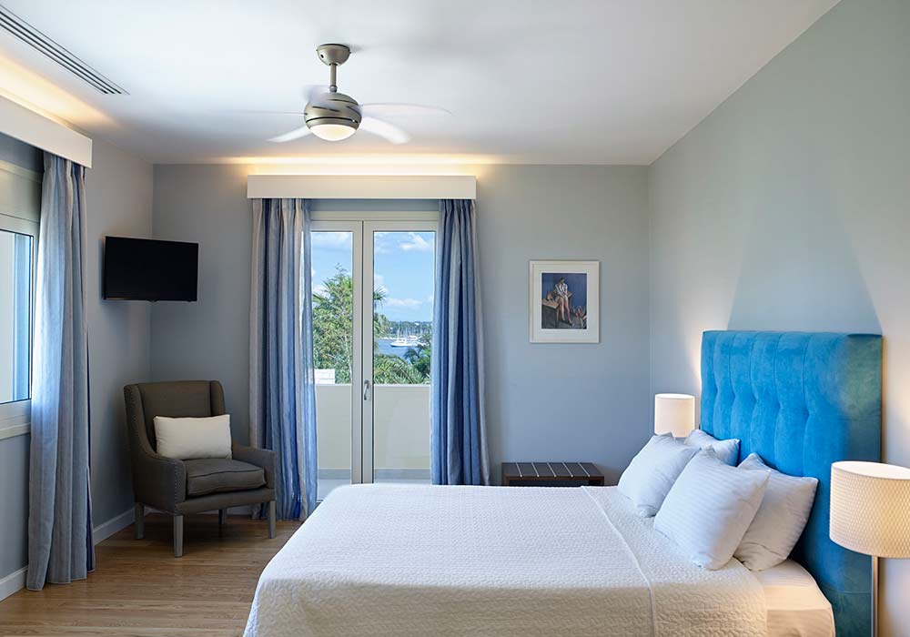 A Villa Alpha bedroom with a blue headboard, ceiling fan and doors open to a balcony over the trees