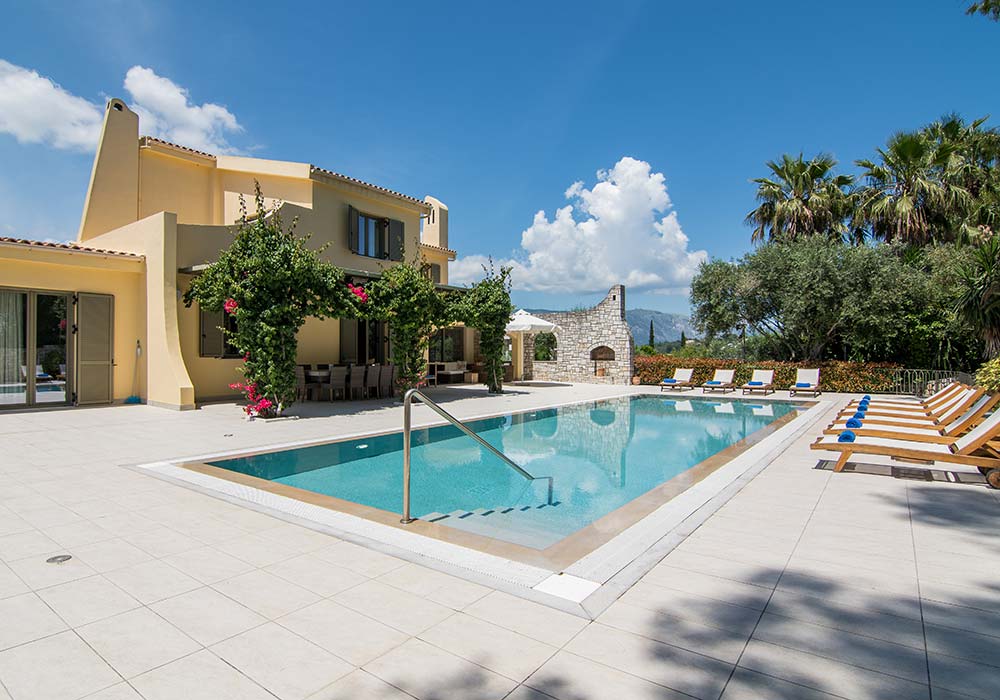 Villa Alpha's pool from the terrace, loungers along one side, palms and a stone garden wall beyond